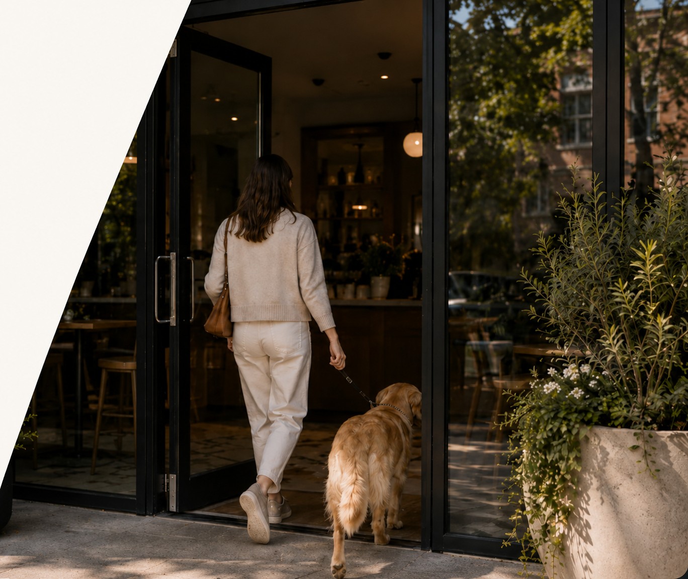A woman and her golden retriever walking through the open glass door of a café, greenery and a stone planter beside the entrance