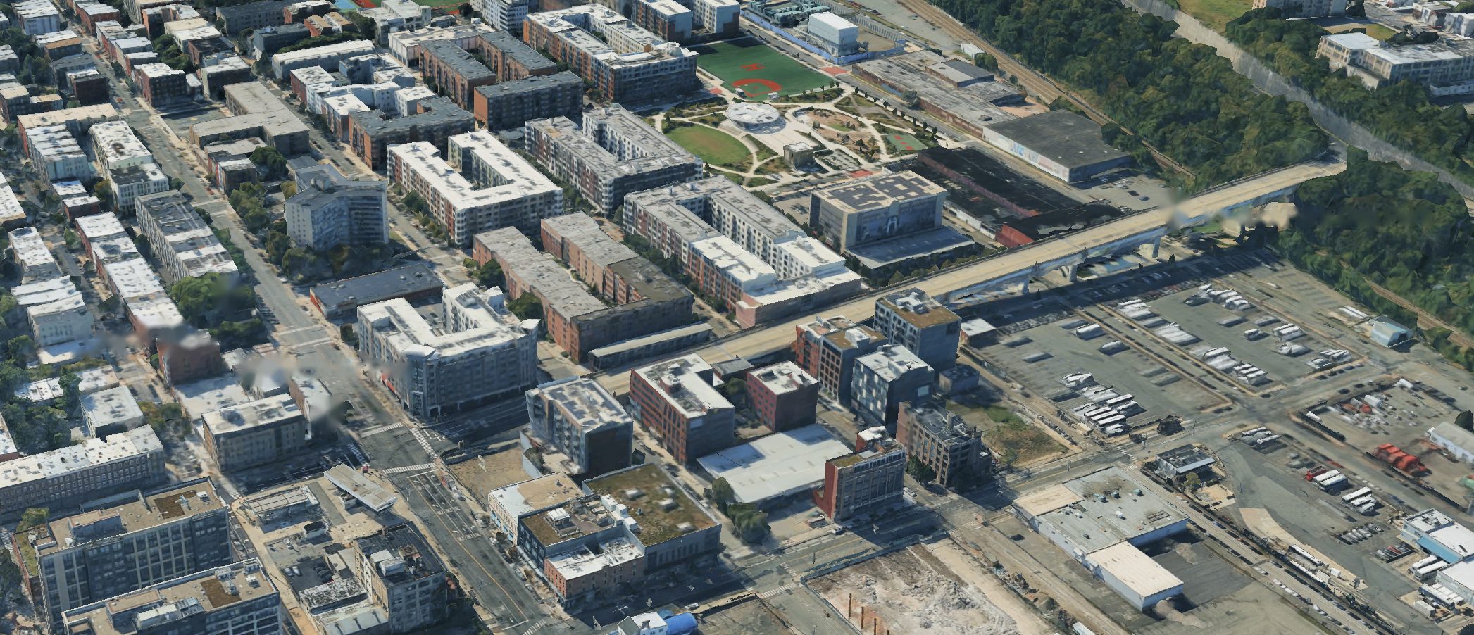 An aerial view of a city neighbourhood: apartment buildings along a busy road, a small tree-filled park, crosswalks at an intersection, an elevated viaduct park with a dog run, and large truck and bus lots