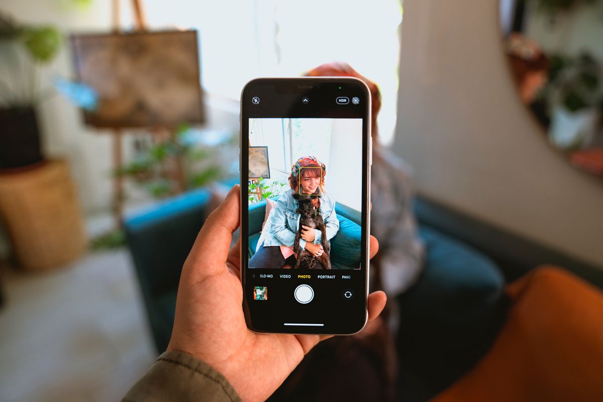 A hand holding up a phone, taking a photo of a woman sitting with her dog on a sofa