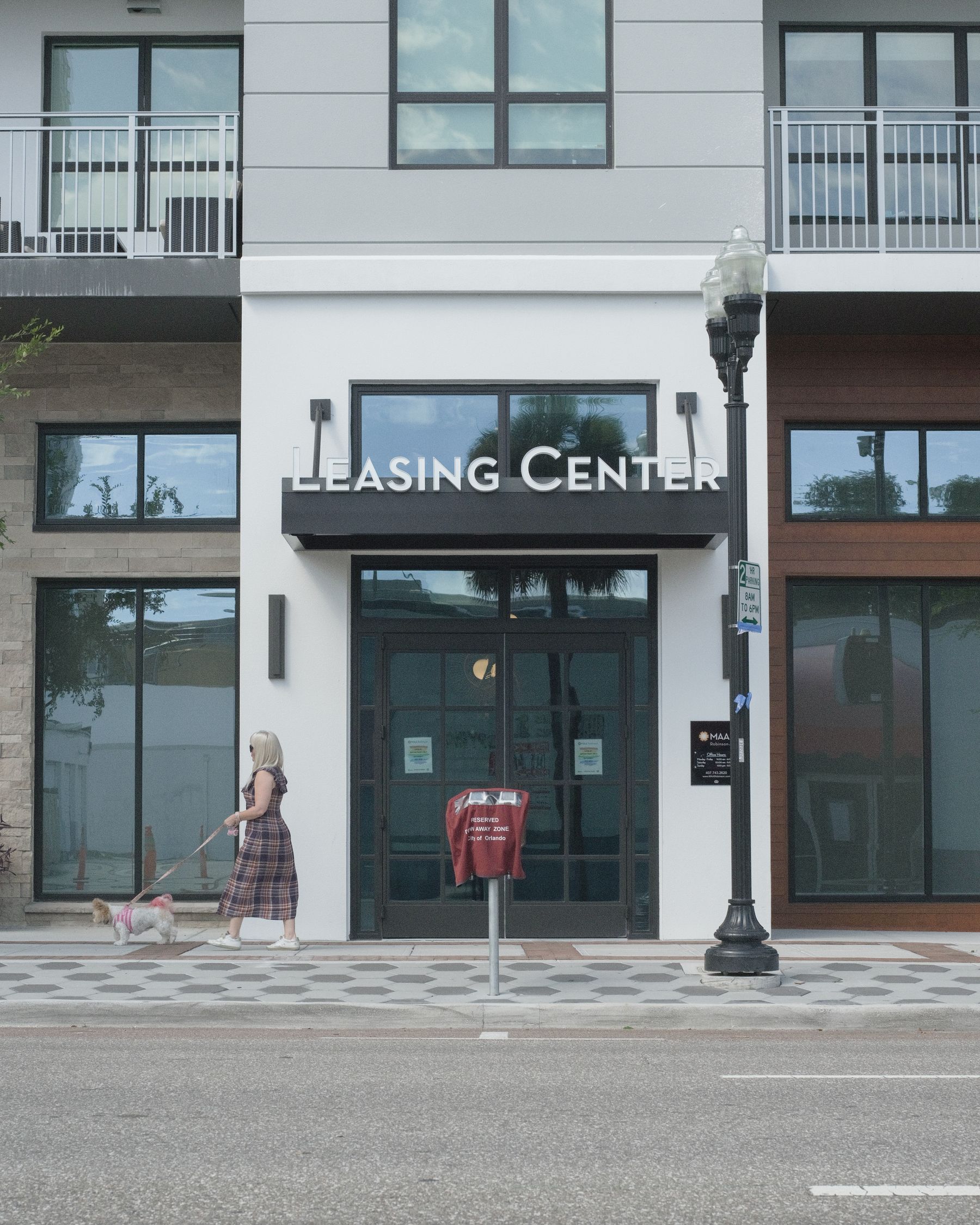 The street entrance of an apartment building's leasing center, with a woman walking a small dog past the door